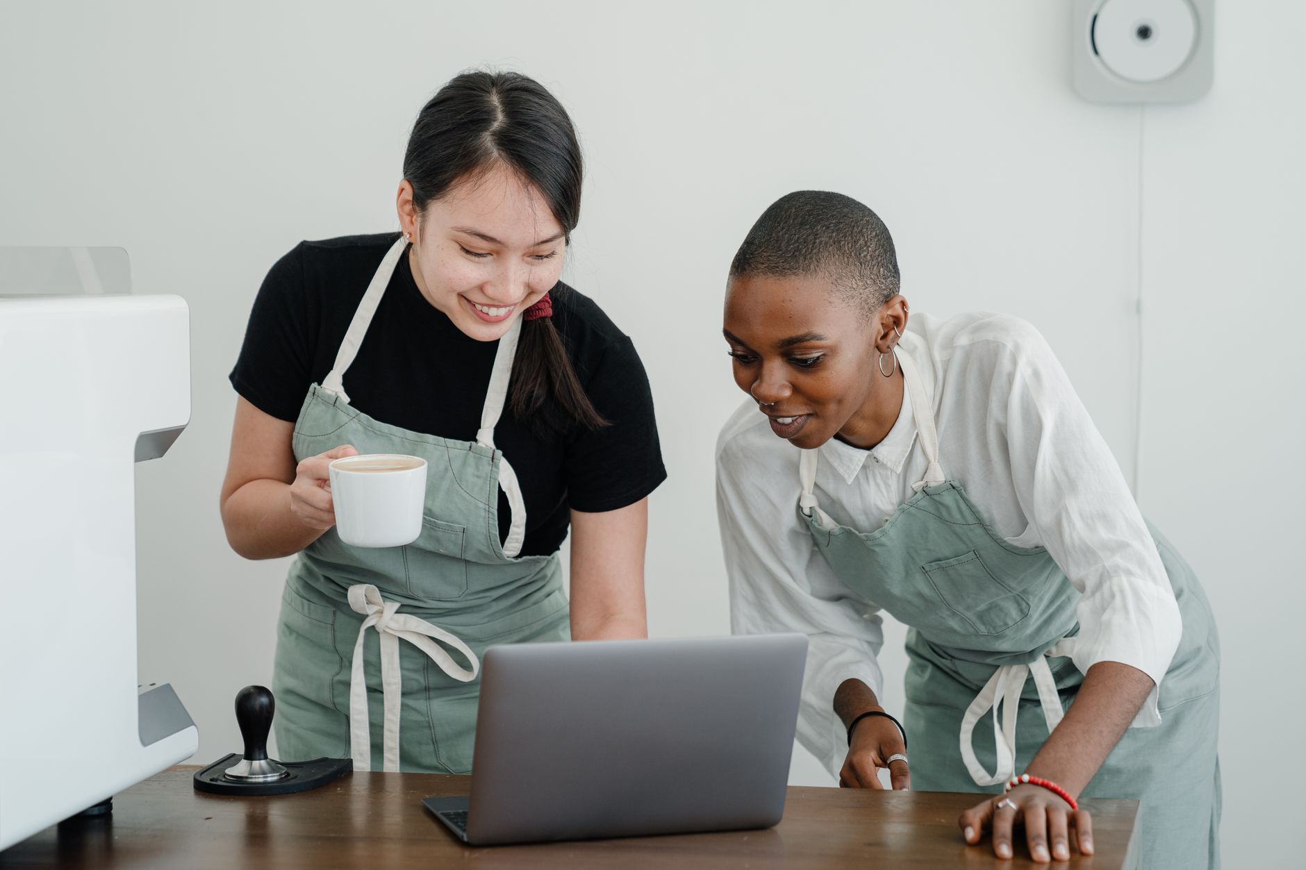 Online Barista Eğitimi Nereden Alınır? 1 Positive young Asian and African American female baristas in aprons smiling and looking at laptop screen while standing near table and enjoying break at work