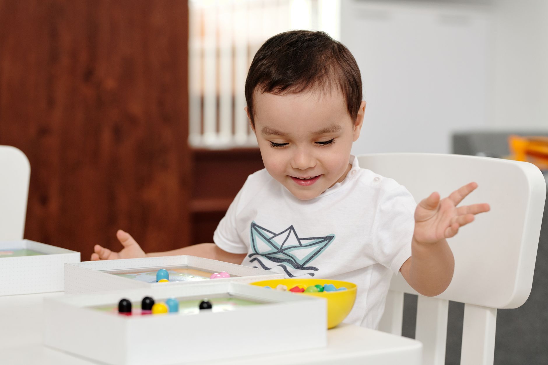 OYUN TERAPİSİ EĞİTMEN EĞİTİMİ Sertifikası 1 A cheerful child engaged in play with colorful beads indoors, expressing delight and curiosity.