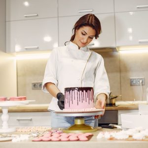 From below of concentrated female pastry cook in uniform garnishing pink cake with marshmallows among confectionery items in kitchen