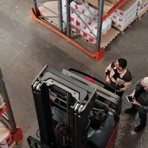 Two workers in a warehouse discussing logistics near a forklift captured from above.