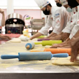 Chefs wearing uniforms and masks prepare dough with colorful rolling pins in a professional kitchen.