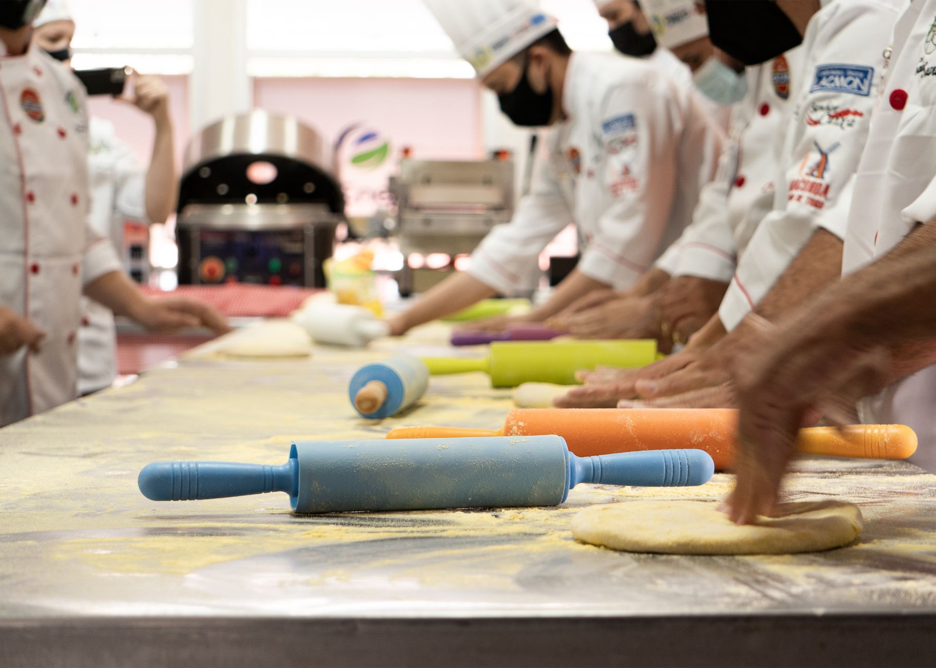 PROFESYONEL AŞÇILIK EĞİTİMİ Sertifikası 1 Chefs wearing uniforms and masks prepare dough with colorful rolling pins in a professional kitchen.