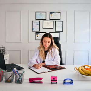 Nutritionist sits at a modern office desk writing, with fruits and diplomas displayed, emphasizing health and professionalism.