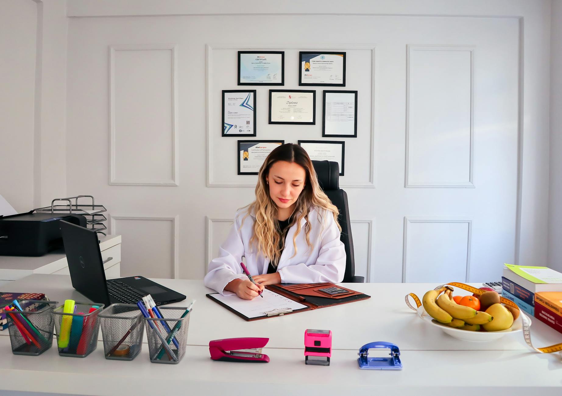 YETİŞKİN ODAKLI TERAPİ EĞİTİMİ Sertifikası 1 Nutritionist sits at a modern office desk writing, with fruits and diplomas displayed, emphasizing health and professionalism.