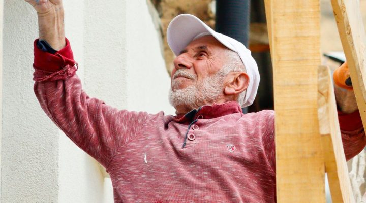 Mantolama Ustalık Belgesi Nasıl Alınır? 12 İskelede oturan şapkalı yaşlı adam dış duvarı boyuyor.
